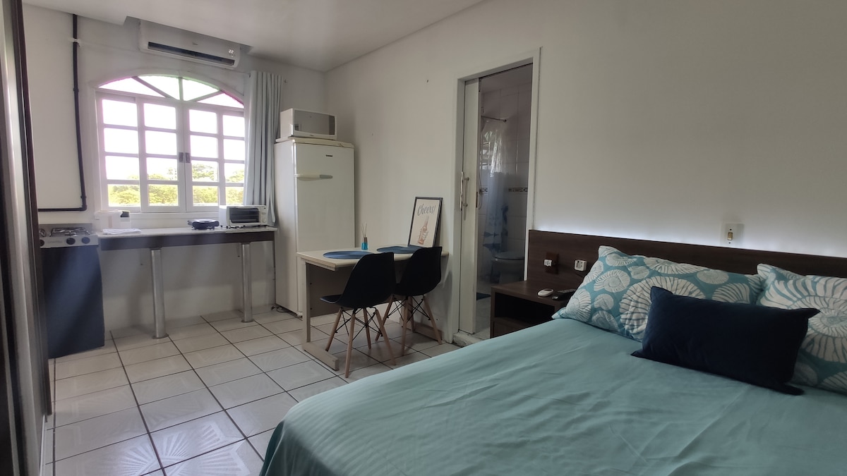 A bright living area features a spacious bed with a teal coverlet and decorative pillows. A compact dining table with black chairs is positioned near a window, illuminating the space. A kitchenette is arranged with a refrigerator and stovetop, enhancing the area’s functionality.
