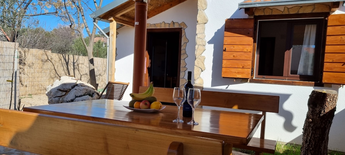 An outdoor dining area features a wooden table set with a bowl of fruit and glasses beside a bottle of wine. The tranquil setting is complemented by a nearby rustic stone house and warm sunlight filtering through the trees.