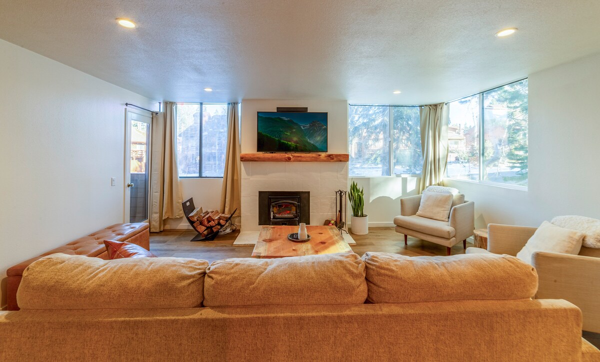 A spacious living area is highlighted by a large, comfortable couch and a modern wood-burning fireplace. Natural light streams through multiple large windows, creating a bright ambiance. A TV is mounted above the fireplace, and minimalistic furniture, including a stylish armchair, complements the setting.