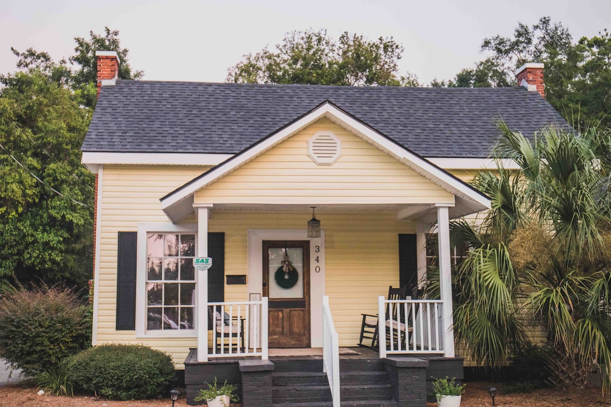 The exterior of a charming yellow house features a welcoming front porch with white railings and rocking chairs. Lush greenery and palm plants surround the entrance, while the front door is elegantly framed by a decorative wreath. A gentle slope leads up to the porch steps.