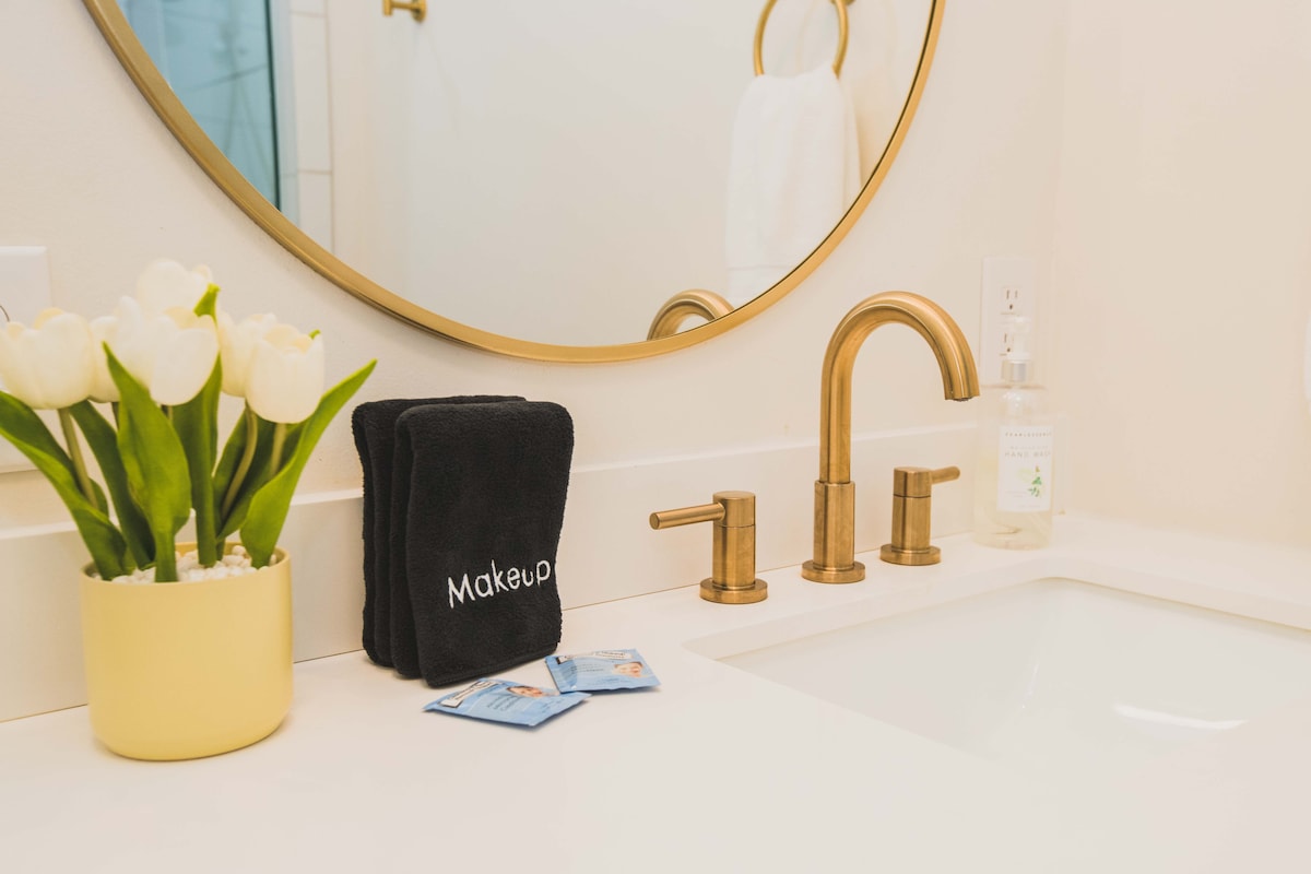 A bathroom sink area is presented, featuring a circular mirror above a white countertop. A modern faucet with bronze finishes is installed, alongside a bottle of hand soap. A potted plant with white tulips adds a touch of greenery, while a black towel labeled 'Makeup' rests nearby.