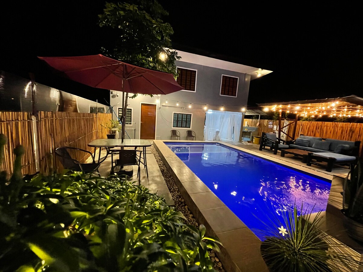 The outdoor area features a shimmering pool under a starry sky, framed by warm lighting. Nearby seating arrangements include a shaded dining table and lounge chairs, providing a welcoming space for relaxation. The house, with its inviting windows, complements the tranquil atmosphere.