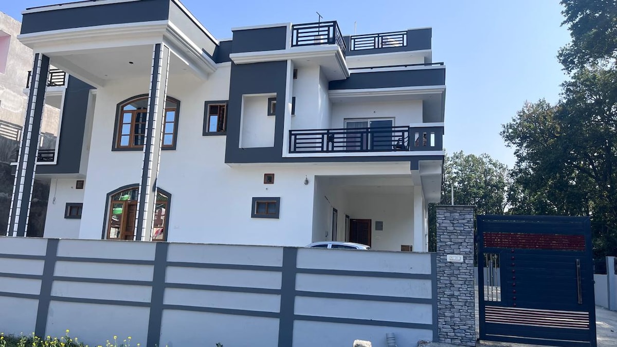 A modern two-story building features a sleek exterior of white and gray tones. The design incorporates multiple balconies and large windows, promoting natural light. A stone wall surrounds the property, providing privacy. A well-maintained entrance gate is visible at the front.