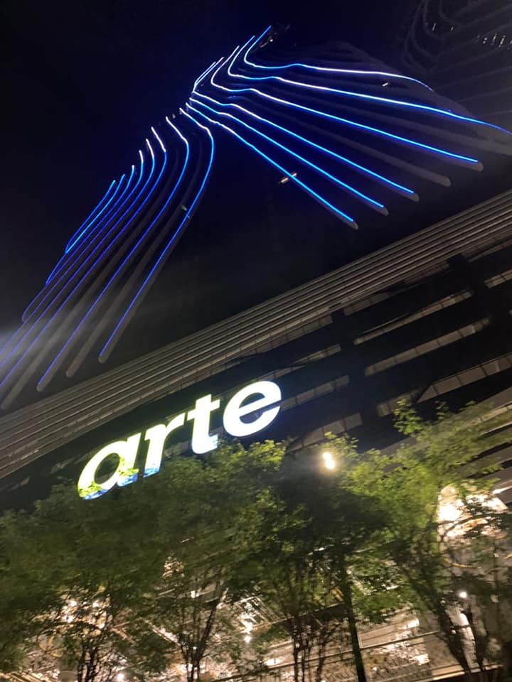 The building's exterior is illuminated with striking blue lights, creating a vibrant visual presence against the night sky. The prominent 'arte' sign is featured at the base, surrounded by lush greenery that enhances the urban setting.