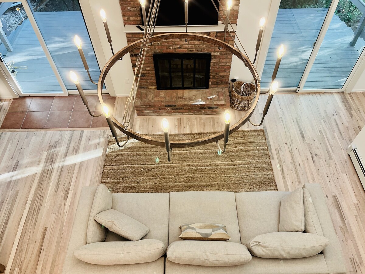 A spacious living area is highlighted by a large chandelier hanging above a light-colored oversized couch. A textured area rug lies beneath, complementing the wooden flooring. A brick fireplace is located in the background, while large glass doors offer views of the outdoor deck.