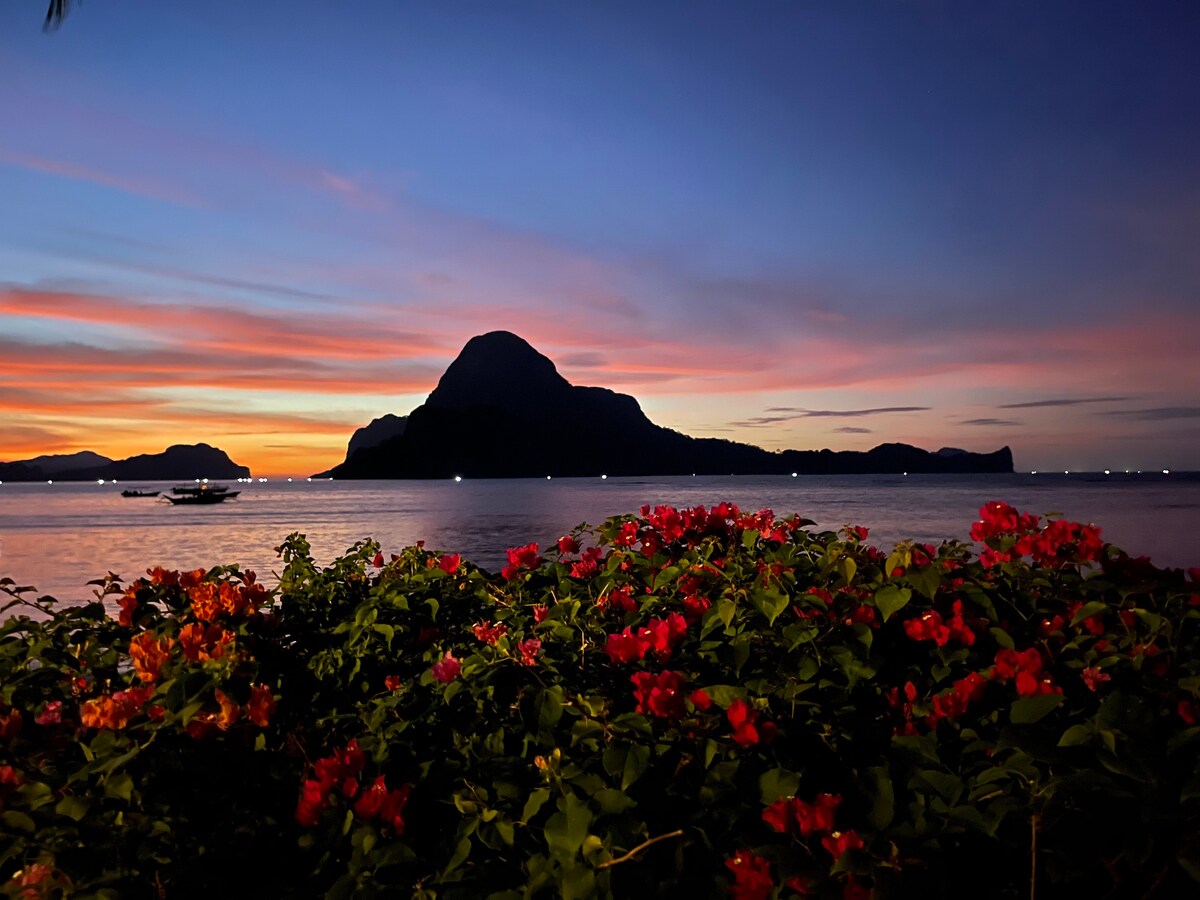 A vibrant scene is depicted with blooming flowers in shades of red and orange in the foreground, while a tranquil water view stretches out to a mountain silhouette against a colorful sunset sky. Soft, fading light reflects on the calm water surface.