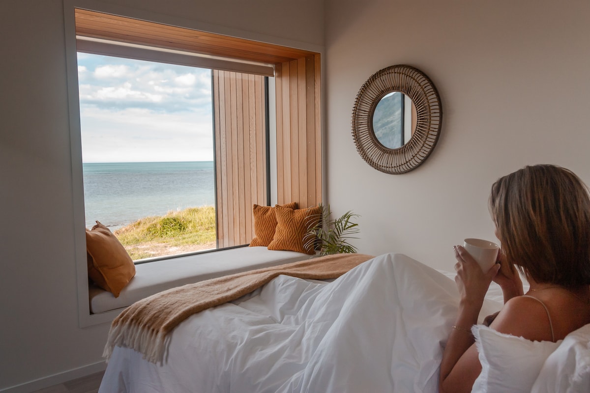 A cozy bedroom features a spacious bed with a soft blanket and fluffy pillows. A large window provides an unobstructed view of the ocean, framed by wooden accents. Natural light fills the space, enhancing the serene atmosphere.