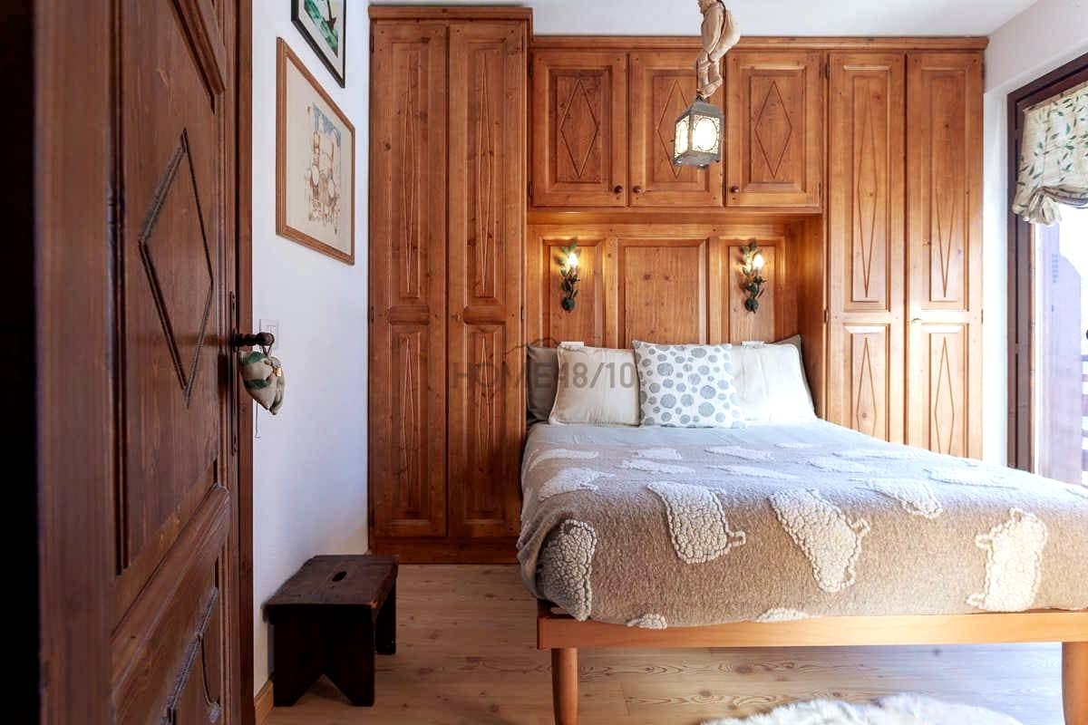 A cozy bedroom features wooden cabinetry that complements the warm tones of the furnishings. A spacious bed with a textured blanket sits against the wall, illuminated by decorative wall sconces. Natural light flows through a window adorned with patterned curtains.