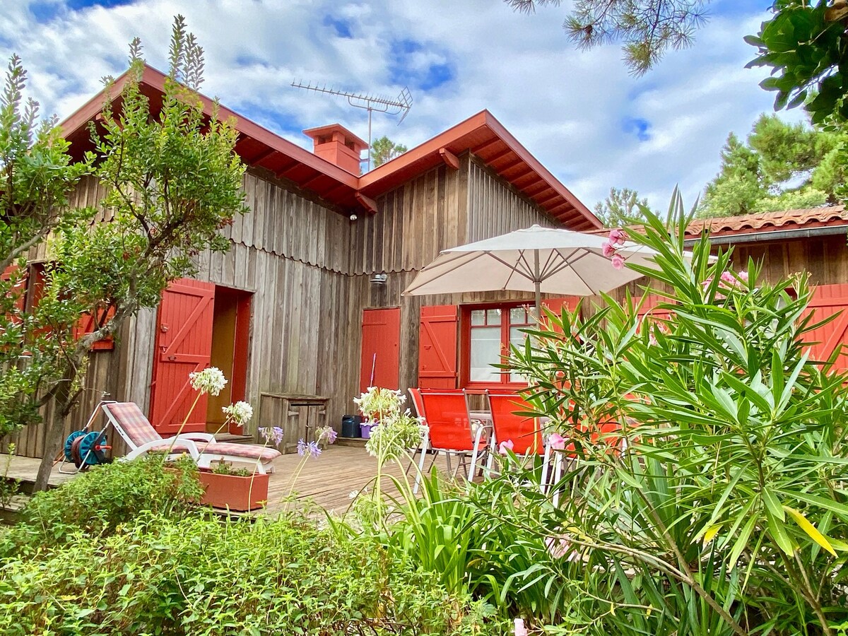 A rustic wooden exterior of the house features bright red shutters and a wooden deck adorned with seating. An umbrella offers shade over a seating area with bright red chairs, surrounded by lush greenery and blooming plants.