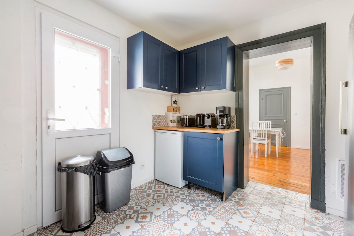 The kitchen area features navy blue cabinets and a countertop with small appliances arranged neatly. A stainless steel trash bin and a black waste bin are positioned beside the cabinetry. Light enters through a window, illuminating the patterned floor tiles.