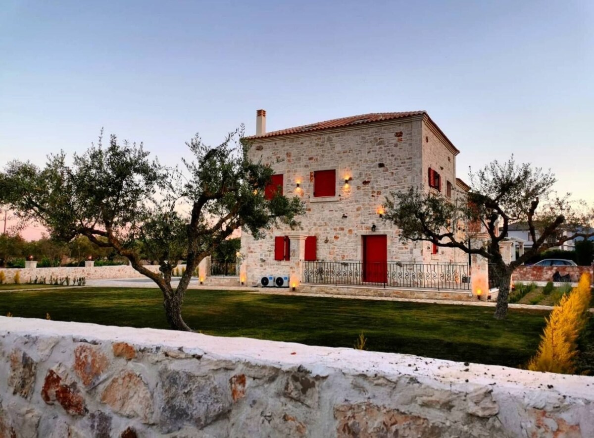 The exterior of the villa is presented, showcasing a rustic stone facade with red shutters and door. The landscaped garden features olive trees and manicured grass, bordered by a white stone wall. Subtle lighting enhances the villa's charm as twilight sets in.