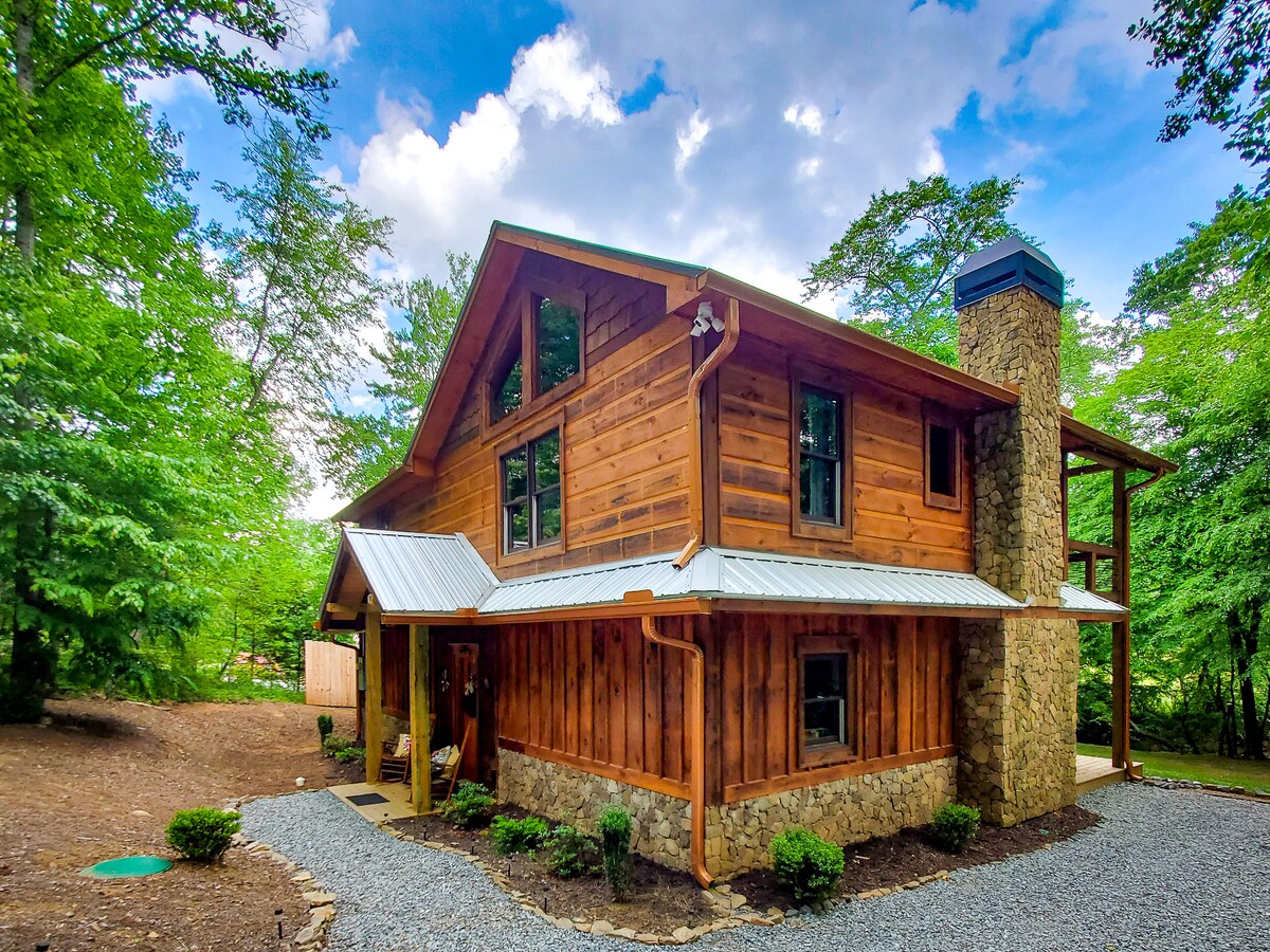 The exterior of the cabin features rich wooden siding and a stone chimney, surrounded by lush greenery. A steeply pitched roof is adorned with a metal overhang, while large windows invite natural light. The gravel driveway complements the rustic charm of the retreat.