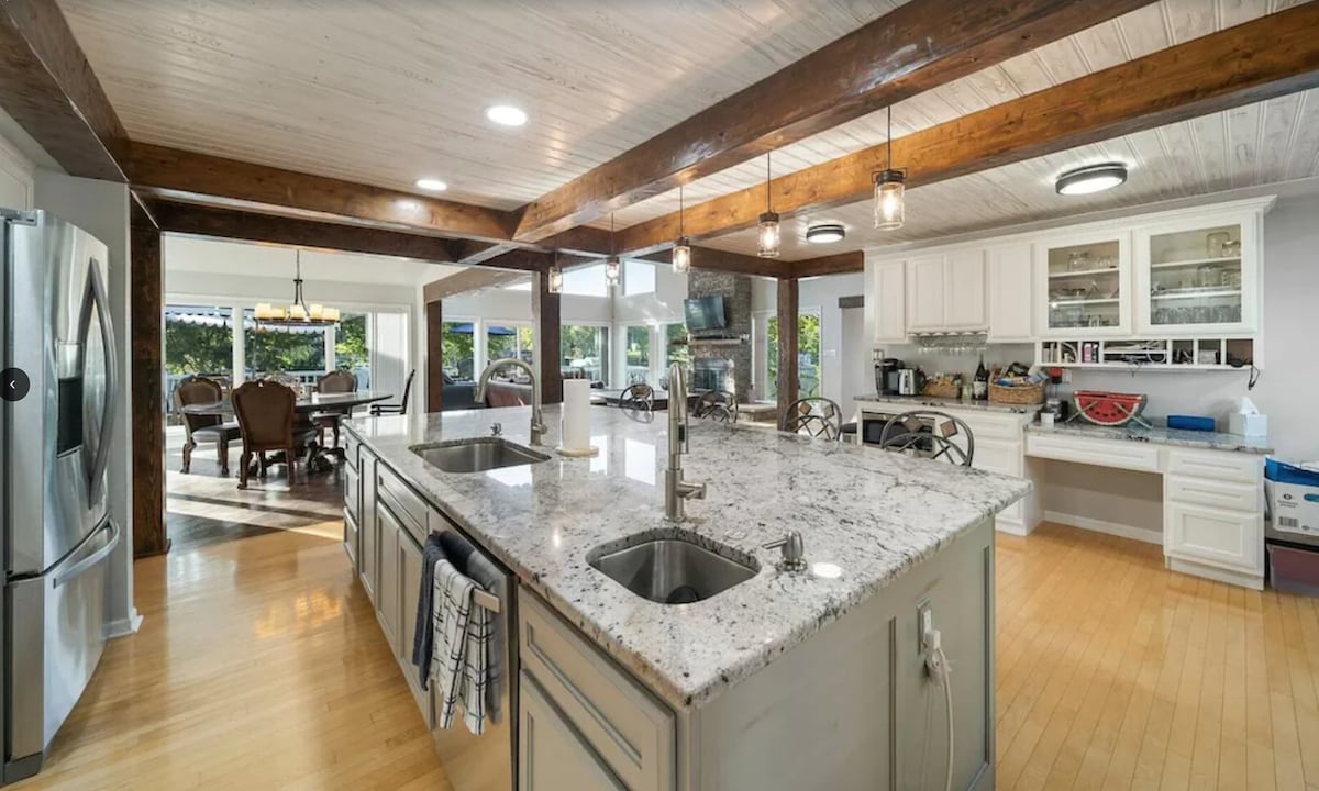 The kitchen features a large granite island with dual sinks, surrounded by wooden beams. Modern appliances, including a refrigerator and coffee station, are visible. Natural light flows through ample windows, illuminating the dining area and creating an open atmosphere.