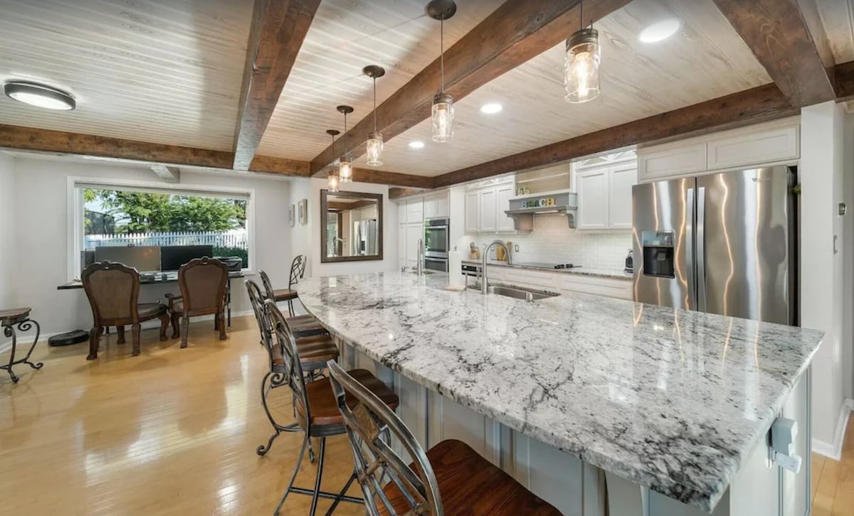 A spacious kitchen features a large granite island with seating and overhead pendant lighting. Stainless steel appliances, including a refrigerator and stovetop, are integrated into white cabinetry. A dining area with elegant chairs is visible in the background, illuminated by natural light from nearby windows.