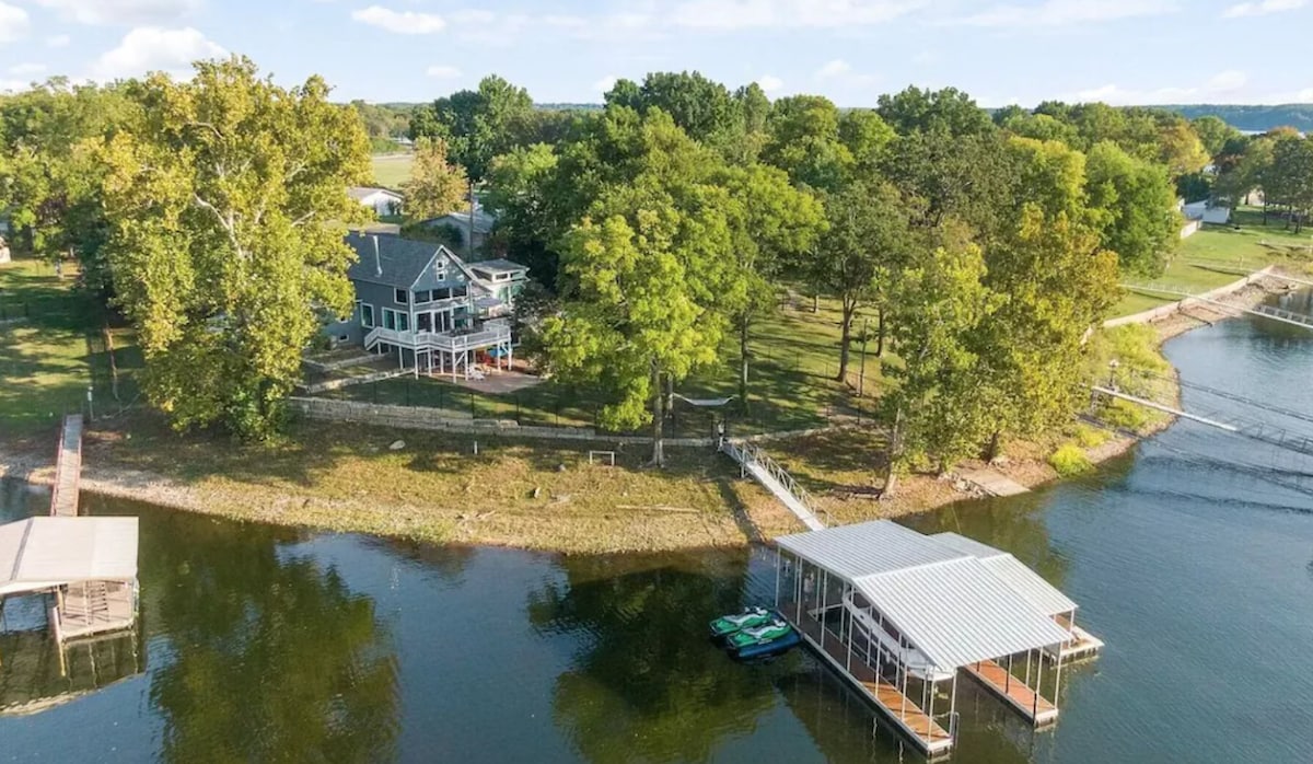An aerial view showcases a serene waterfront property surrounded by lush greenery. A private dock is visible, with a boat lift and recreational equipment nearby. The house features multiple outdoor spaces, nestled comfortably against the backdrop of trees and the gentle water.