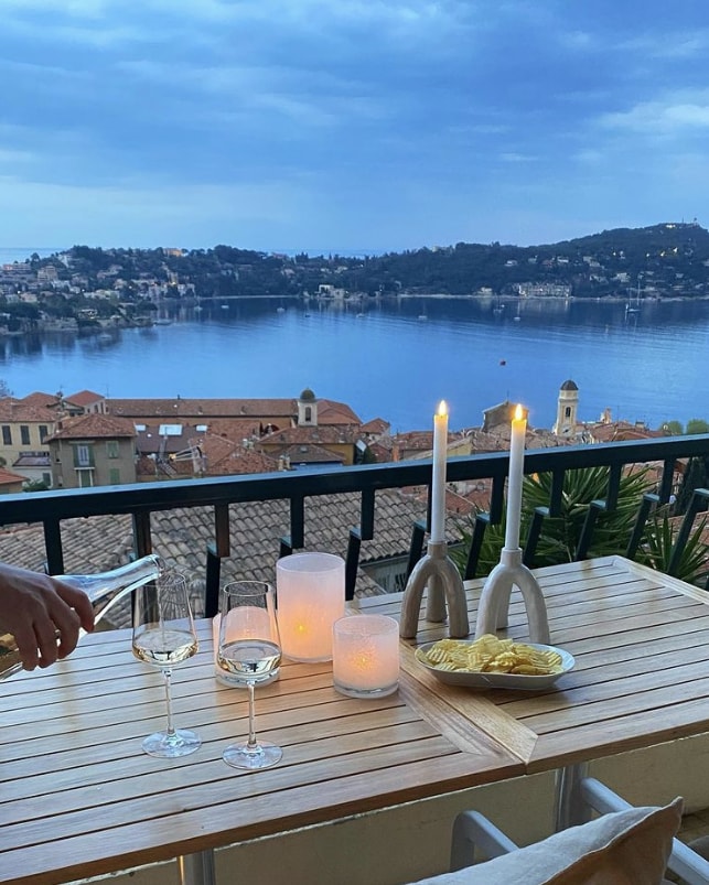 A balcony table is set with candles, glasses of white wine, and a plate of snacks, creating an inviting atmosphere. The view showcases the tranquil bay, with rooftops of Villefranche-sur-Mer and hills in the background, under a softly lit sky.
