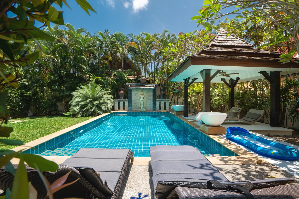 A tranquil pool area is highlighted with a shimmering blue pool, surrounded by lush tropical plants. Comfortable lounge chairs are positioned at the poolside, while a shaded pavilion features seating for relaxation. A fountain adds a serene touch in the background.