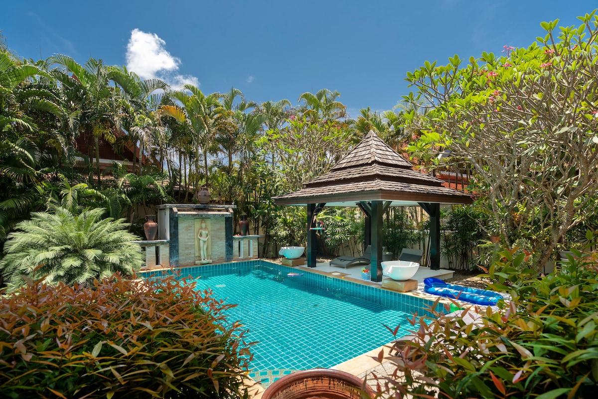 A tranquil pool with clear blue water is surrounded by lush tropical landscaping. A gazebo with a thatched roof offers shaded seating, while vibrant plants enhance the natural setting, creating a serene outdoor space ideal for relaxation.