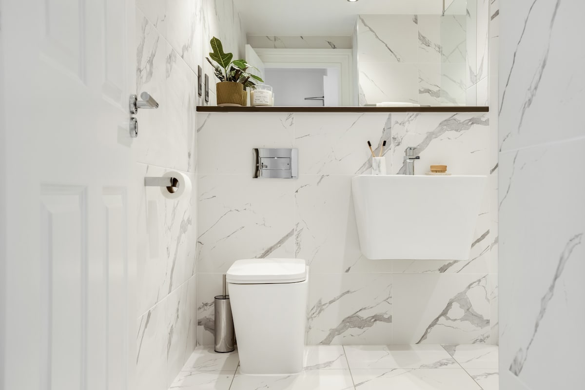 A modern bathroom features white marble walls and floor tiles, creating a clean and bright space. A compact white toilet is positioned next to a minimalistic sink with a wooden shelf above, displaying potted plants and toiletries. Soft lighting enhances the spacious feel.