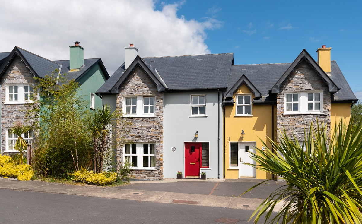 A row of colorful houses is visible, featuring stone and clapboard facades. The house in the foreground has a red door, with light blue and yellow accents on either side. Lush greenery and flowering plants complement the entrance, under a bright blue sky.
