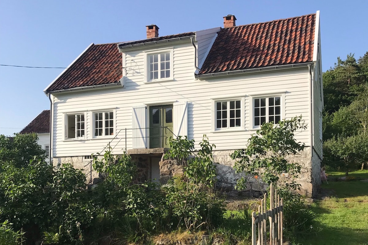 The white house features a charming facade with a distinct red roof and multiple windows, allowing ample natural light. The entrance is highlighted by a small staircase. Surrounding greenery includes low bushes and a well-maintained garden area, contributing to the property's inviting appeal.