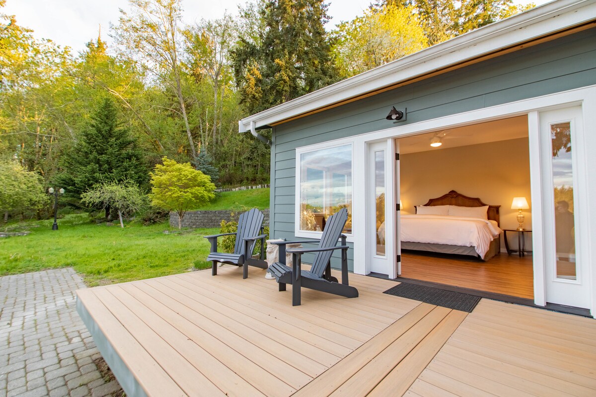 A deck extends from the house, featuring two black Adirondack chairs. Beyond, the spacious interior is visible, displaying a large bed and natural light flooding through expansive glass doors. Lush greenery and trees surround the property, creating a serene environment.