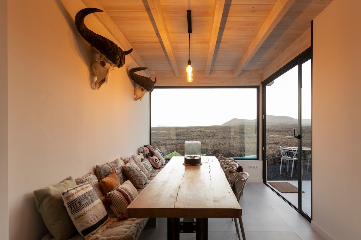 A dining area is set with a long wooden table surrounded by cushioned seating. Two bull skulls are mounted on the wall, accentuating the rustic decor. Floor-to-ceiling glass doors offer views of the volcanic landscape beyond, allowing natural light to fill the space.