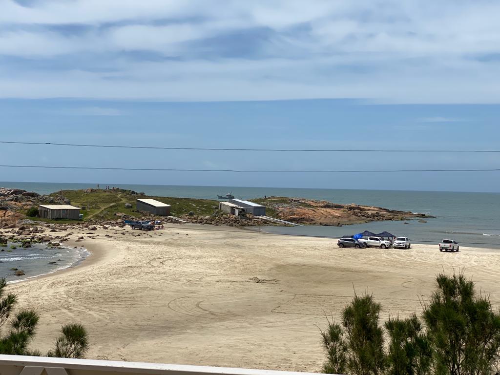 A panoramic view of a sandy beach is displayed, featuring calm waters and a rocky shoreline. Several vehicles are parked along the beach, with minimal activity seen in the distance. Gentle waves are lapping at the shore under a clear blue sky.