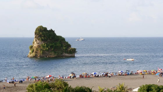 A scenic view of a beach scene captures a rocky island rising from the ocean. Sunbathers and beachgoers occupy the sandy shore, with umbrellas placed for shade. The calm blue waters meet a partly cloudy sky in the backdrop, enhancing the coastal atmosphere.