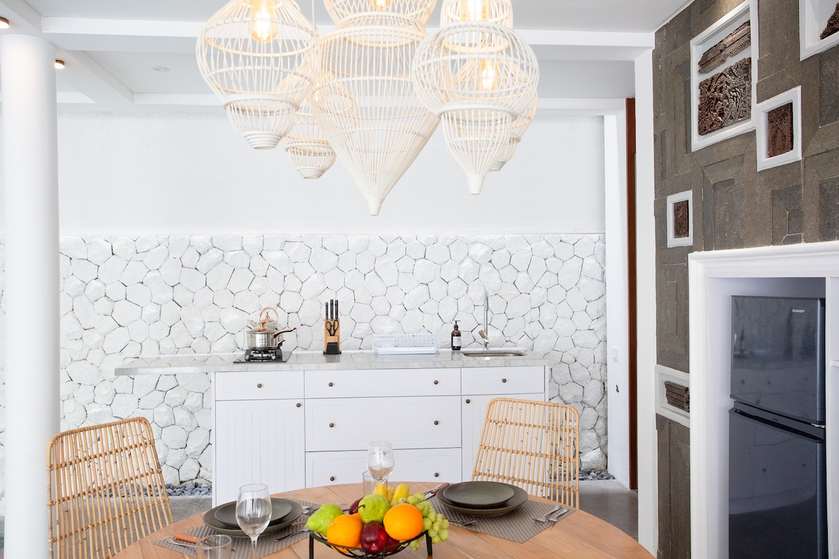 A well-equipped kitchen area features a wooden dining table set for four, complemented by rattan chairs. A unique overhead light fixture adds a natural touch, while a white backsplash, made of small stones, enhances the clean aesthetic. Counter space is utilized with kitchen essentials.