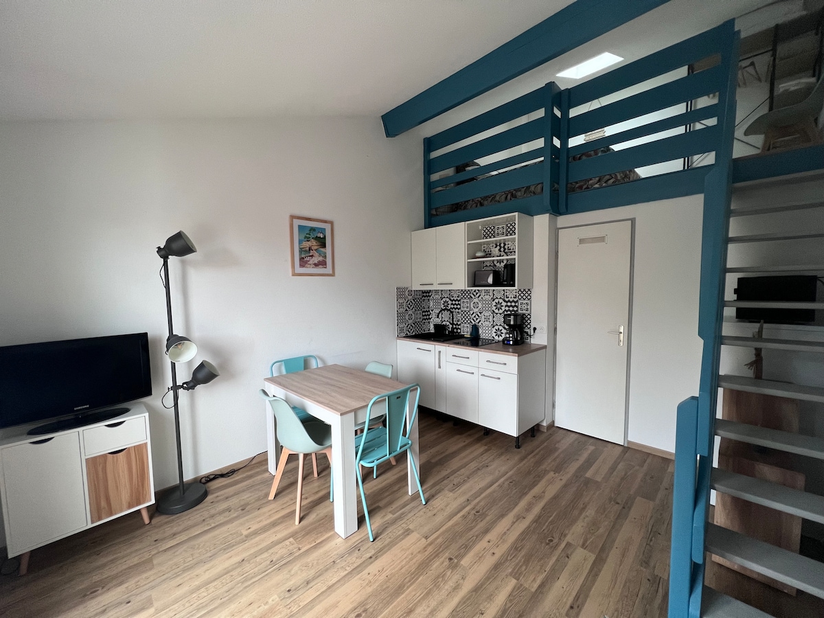 A bright open living area features a dining table with blue chairs, positioned adjacent to a modern kitchen that includes white cabinetry and patterned backsplash. A staircase leads to a mezzanine bedroom above. A television stands on a cabinet next to a floor lamp.