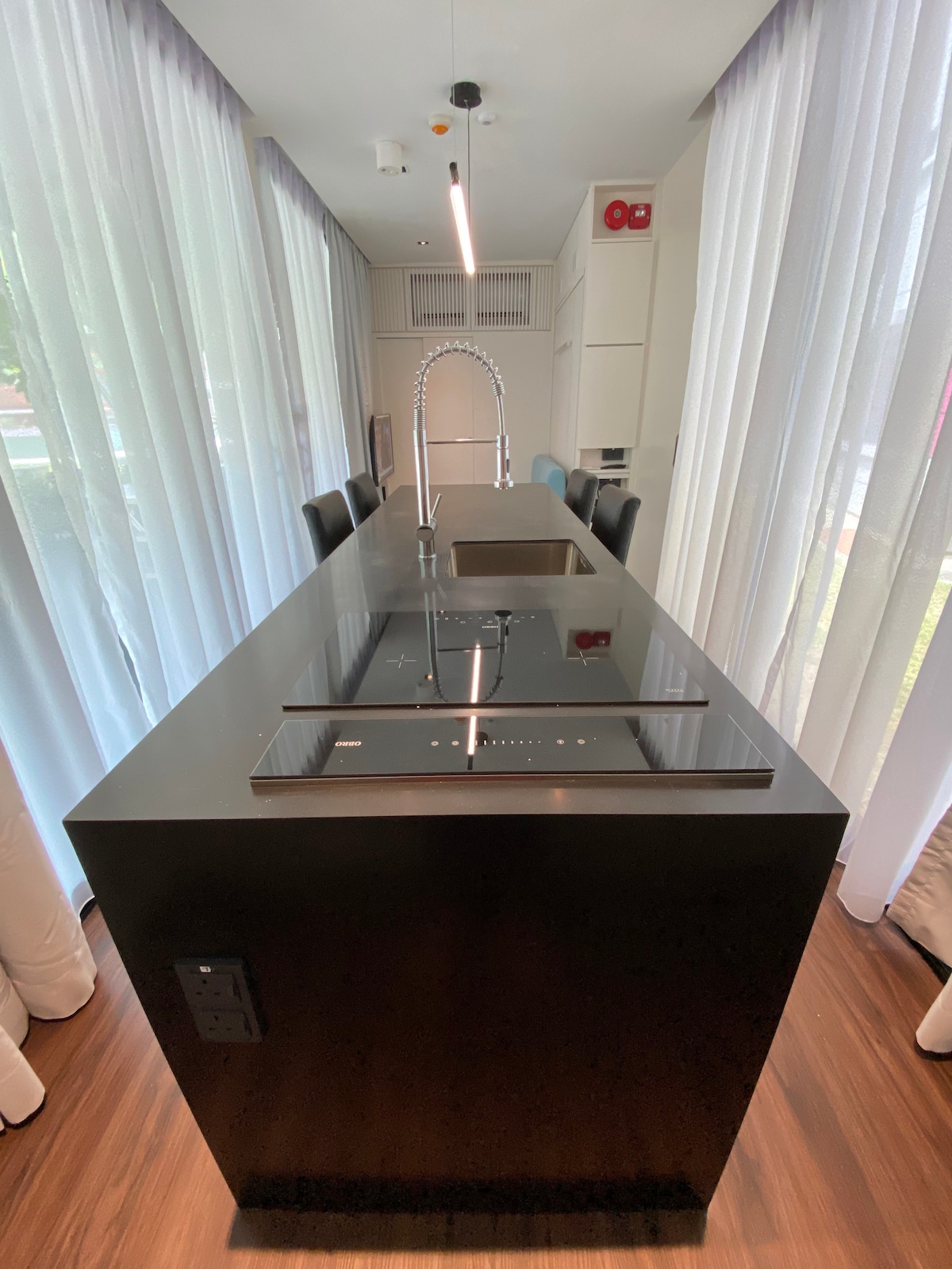 A modern kitchen island features a sleek dark countertop and an integrated cooktop. A polished sink is positioned within the island. Surrounding the island are high-backed bar stools, with large windows dressed in sheer curtains allowing natural light to brighten the space.