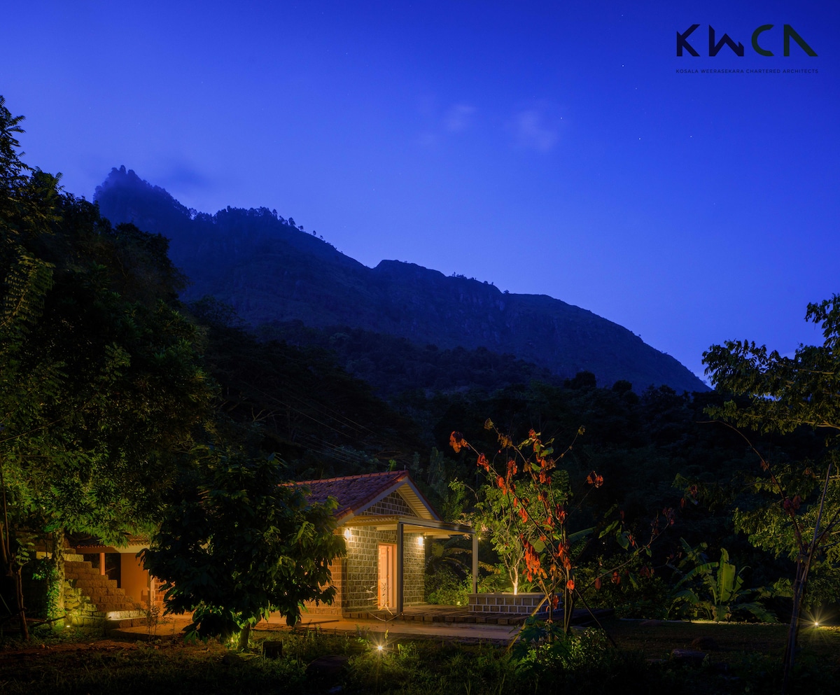 A serene cottage is set against a backdrop of mountains under a twilight sky. Warm light spills from the windows, inviting exploration. Surrounding greenery envelops the space, with trees and flowering plants enhancing the natural beauty of the area.