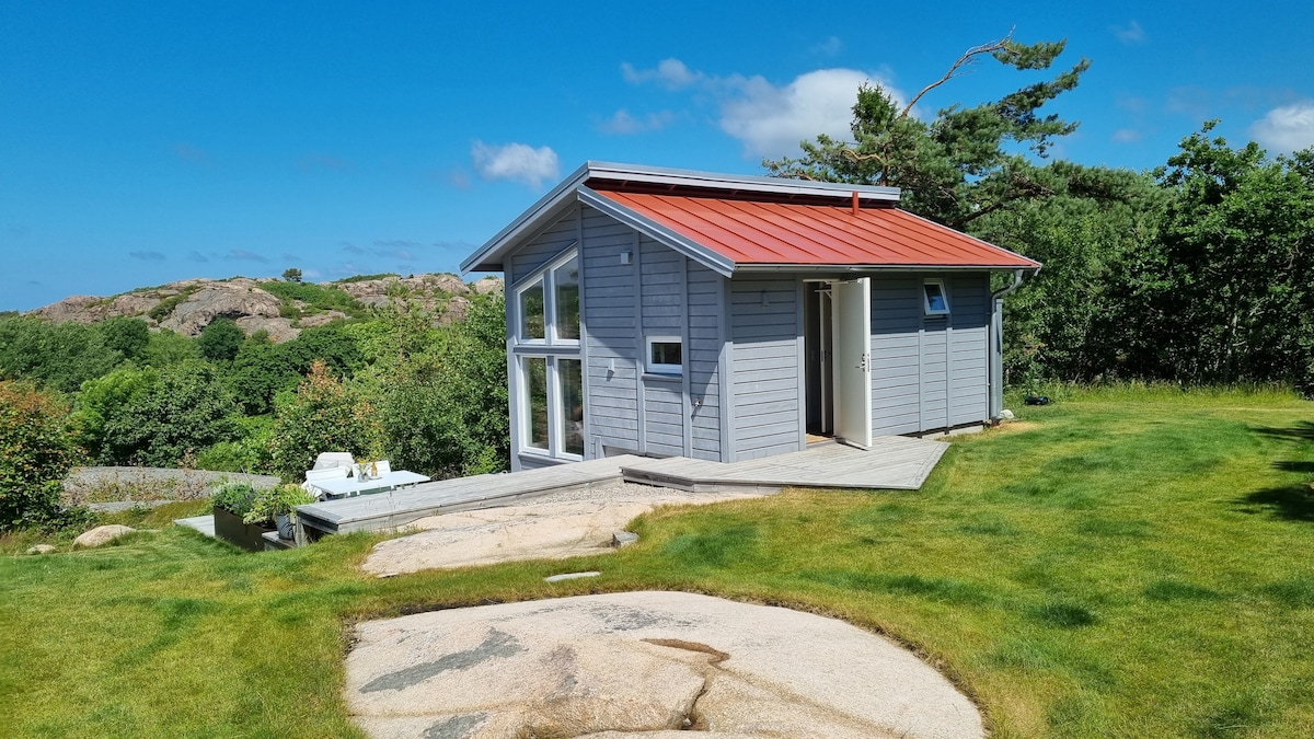 The exterior of a modern, newly built house features large windows, a sloped red roof, and wooden siding. Surrounding greenery and natural rock formations contribute to the serene setting. A wooden terrace is visible, providing an entrance to the house.