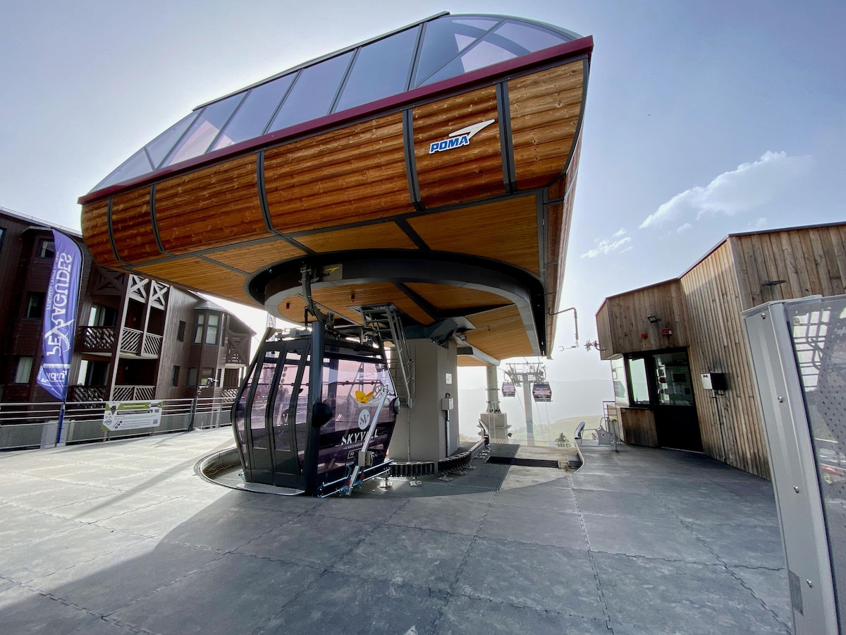 A modern gondola station is featured, elevated on a structured support with wooden accents. The design blends seamlessly with the surrounding architecture. Clear skies and distant mountains are visible, creating a serene setting for visitors embarking on their journey.
