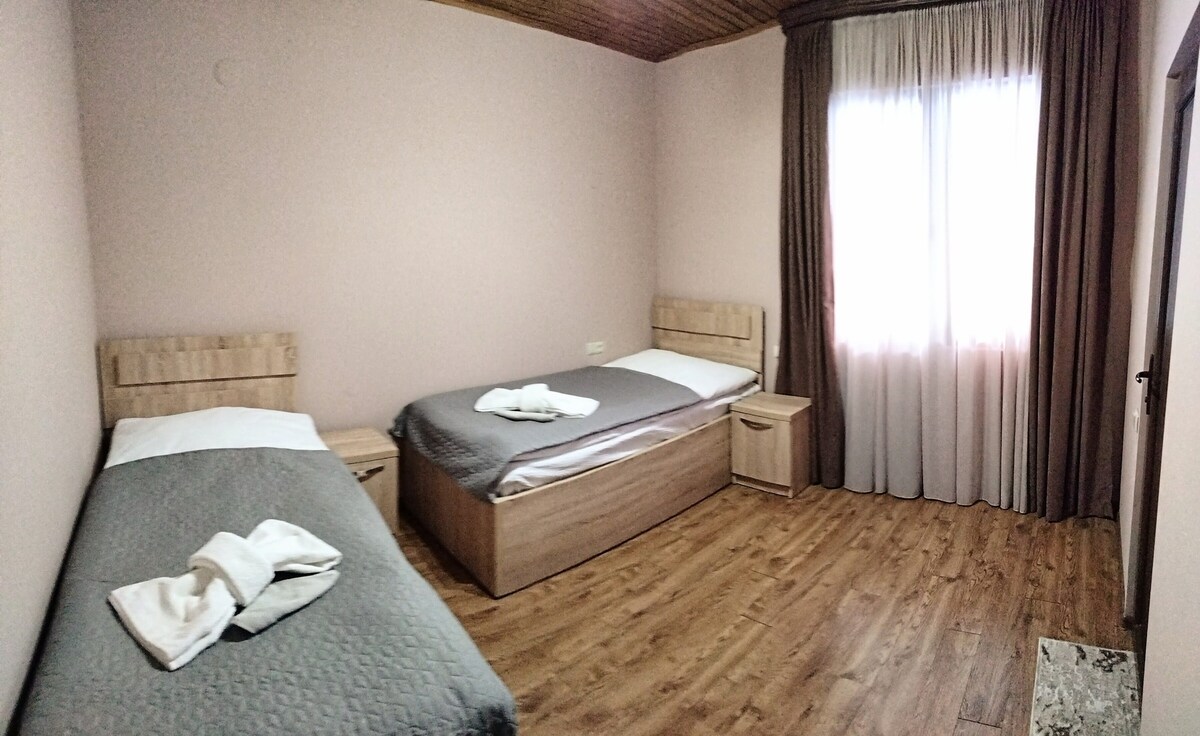 A well-lit room features two separate beds with neutral bedding, complemented by wooden headboards and nightstands. A window dressed with soft, flowing curtains allows natural light to fill the space, while a warm wooden floor adds a touch of warmth to the overall atmosphere.