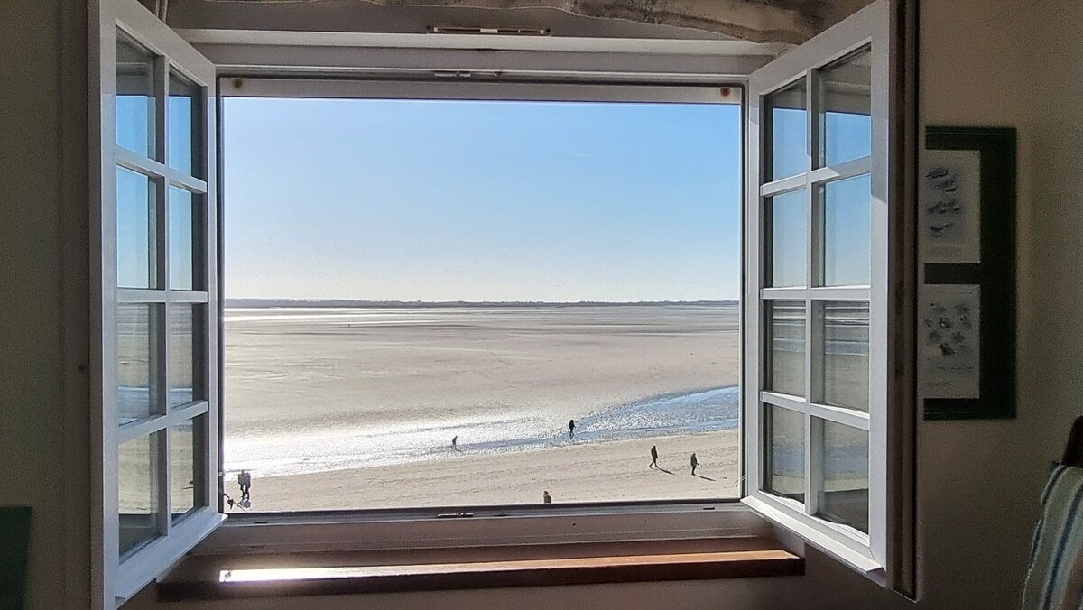 A large window is shown, opened wide to reveal a view of the beach. People can be seen walking along the sandy shore under a clear sky, with the shimmering water reflecting sunlight.