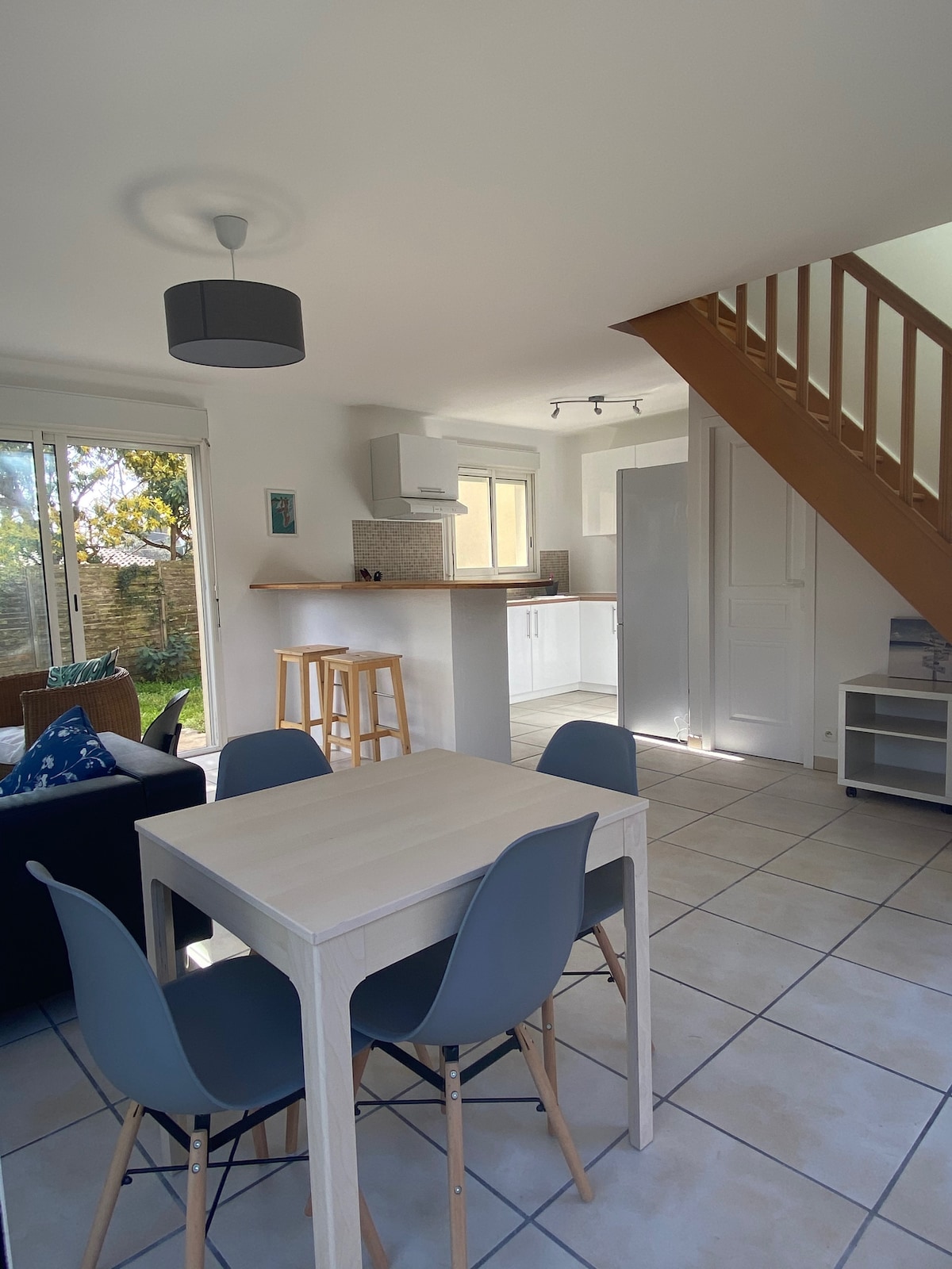An open living space is presented, featuring a light-coloured dining table with four grey chairs. A modern kitchen area can be seen in the background, equipped with a countertop and necessary appliances. Natural light fills the room from large sliding doors leading to the garden.