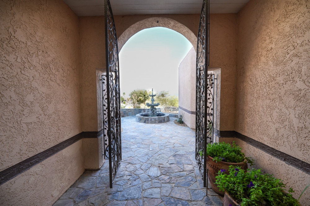 An inviting entrance is framed by ornate, wrought-iron double gates. Natural stone flooring leads to a courtyard featuring a tranquil water fountain surrounded by lush greenery and flowering plants. The open archway offers a glimpse of the serene outdoor setting beyond.