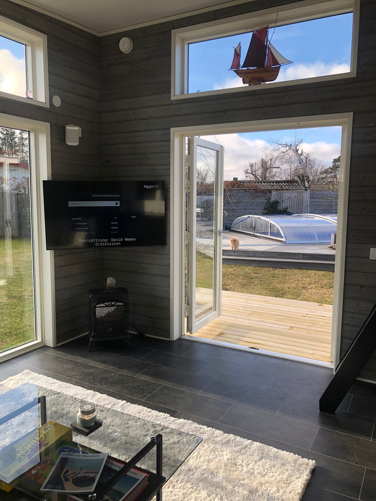 A modern living space is shown with large windows allowing natural light. A television is mounted on a wooden wall opposite a coffee table, with a cozy rug underneath. The view through the open doors reveals a grassy outdoor area and a boat, suggesting a connection to the surrounding nature.