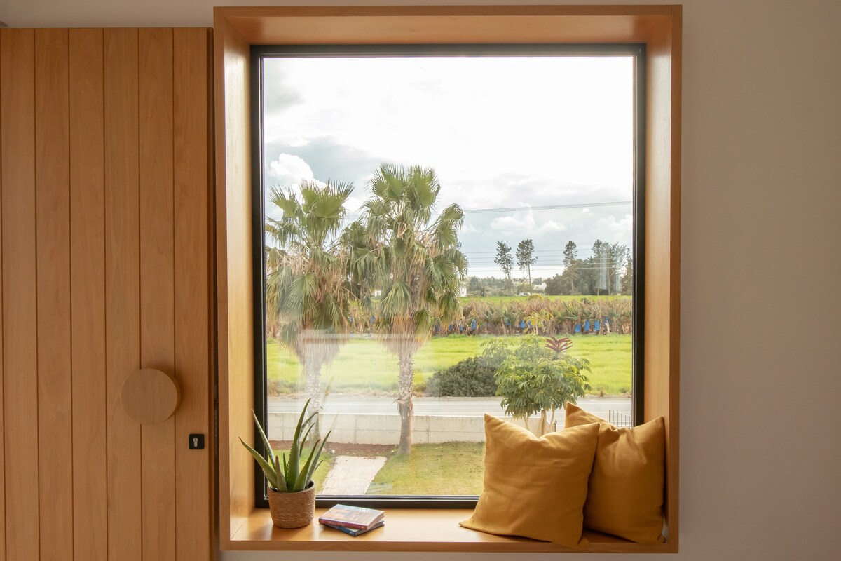 A large window offers a view of greenery and palm trees outside. A wooden ledge is adorned with two decorative pillows and a small plant. A book rests on the ledge, contributing to a sense of relaxation.