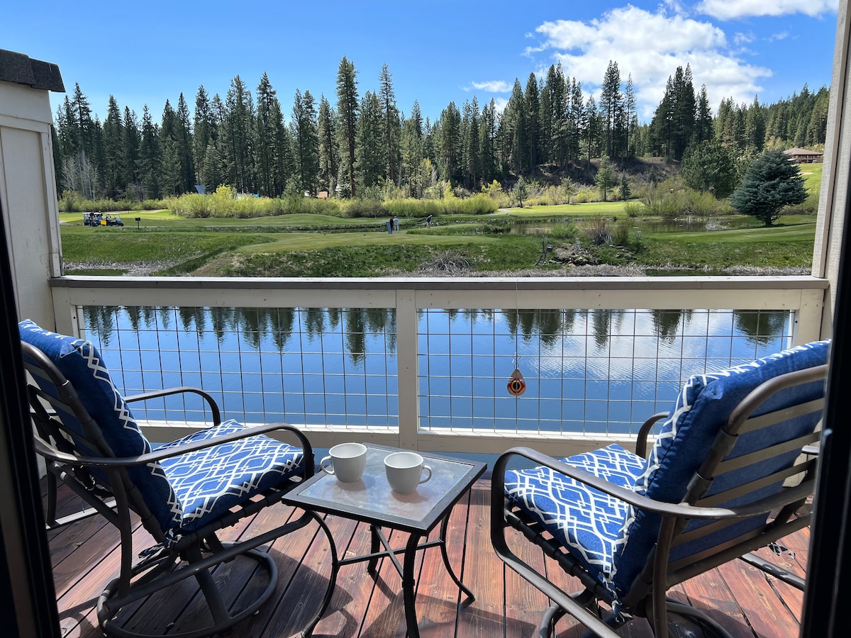 A serene deck overlooks the calm water of Plumas Pines pond, flanked by lush green trees. Two cushioned chairs are positioned around a small table, with two white cups placed on top, inviting moments of relaxation by the tranquil setting.