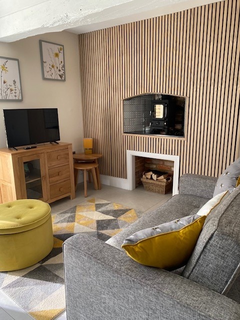 The lounge is accented by a textured wood-paneled wall and features a log burner within a stylish fireplace. A cozy gray sofa sits beside a round ottoman and a small wooden television stand, complemented by a geometric area rug.