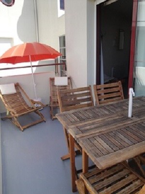 A balcony area is furnished with a wooden dining table accompanied by chairs. Two lounge chairs provide a place for relaxation, and a large red umbrella offers shade from the sun. The setting promotes outdoor dining and leisure.