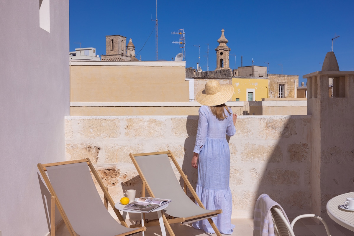 A serene rooftop terrace is presented, featuring two wooden lounge chairs positioned beneath a clear blue sky. A view of historic rooftops and steeples is visible in the background. A figure in a light dress stands, adding a sense of tranquility to the scene.