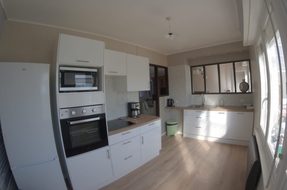 A modern kitchen features sleek white cabinetry and countertops, with built-in appliances including a microwave and oven. Natural light floods the space through a large window, illuminating the wood-look flooring. The kitchen includes a coffee maker and a kettle, contributing to a functional cooking area.