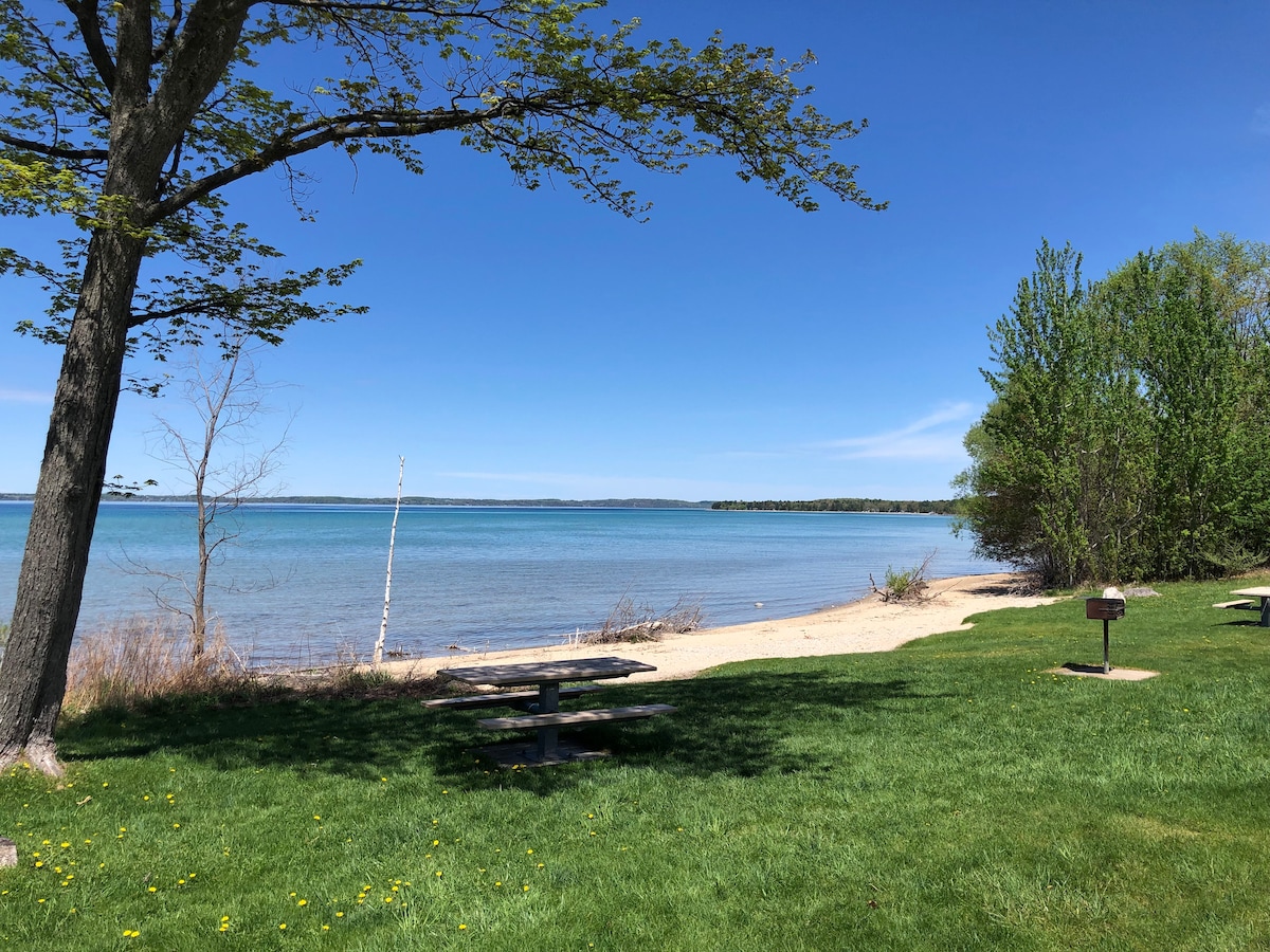 A serene waterfront scene captures the calm waters of East Bay, framed by trees and a sandy stretch of beach. Picnic tables are positioned on the lush green grass, offering a space to enjoy the view. Gentle waves lap at the shoreline under a clear blue sky.