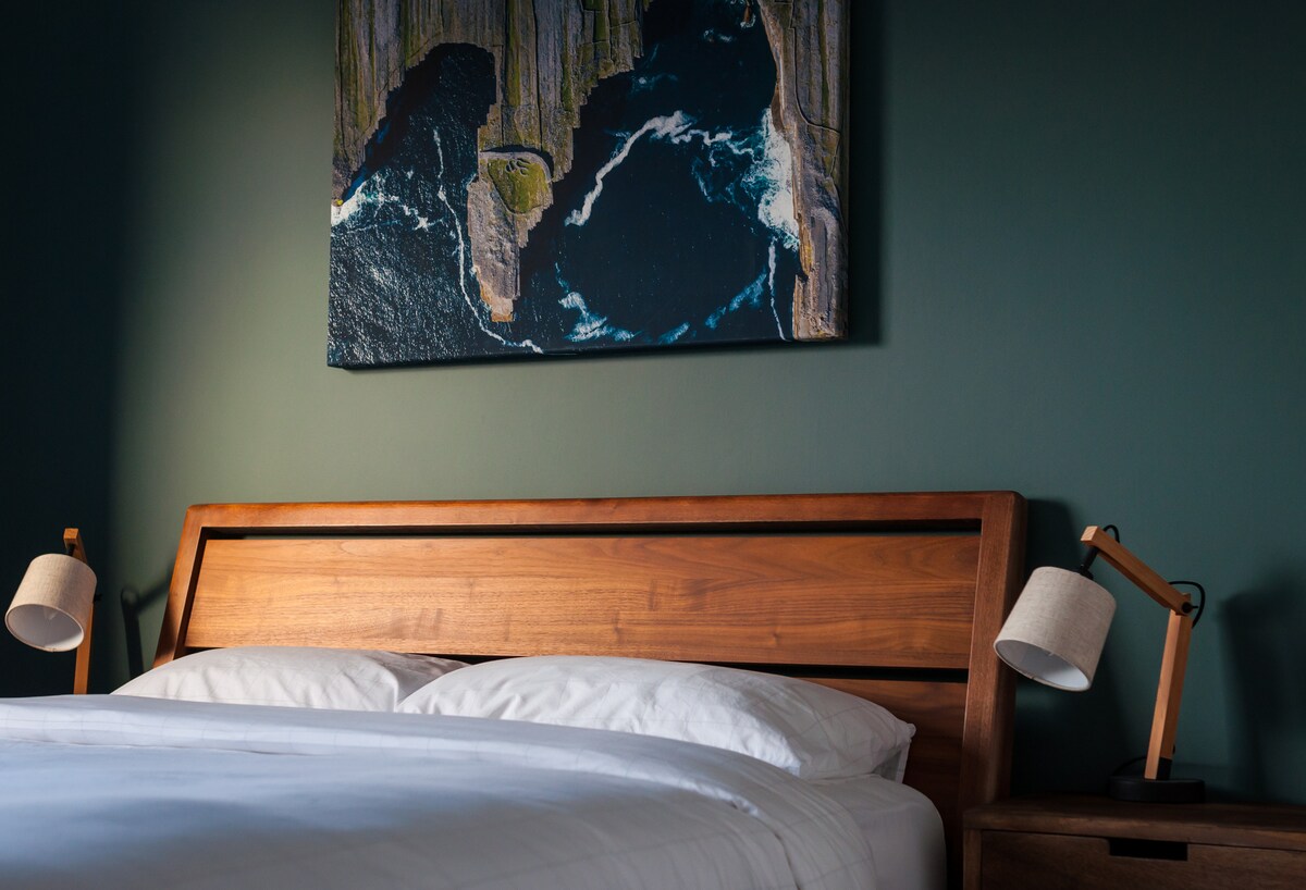 A modern bedroom features a wooden bed frame with a spacious mattress, dressed in crisp white bedding. Soft bedside lamps provide warm light on either side. Above the bed, a landscape painting displays a coastal scene, complementing the deep green wall.