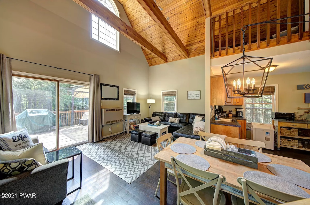 An open-concept living area features a high wooden ceiling and large windows, allowing natural light to fill the space. A comfortable seating arrangement is positioned around a mounted TV, while a dining table with place settings awaits. The adjacent kitchen area is visible in the background.