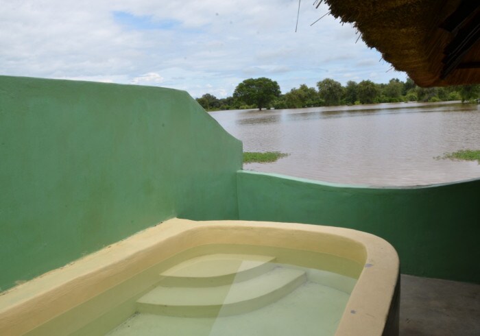 An inviting outdoor space features a large soaking tub with steps, surrounded by green walls. Water reflects the sky and landscape beyond, creating a serene view of the nearby flooded area with trees visible in the distance.